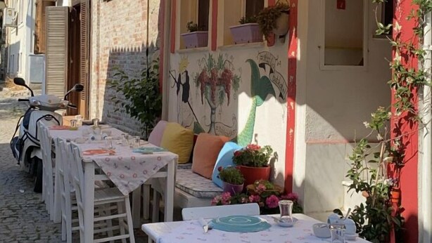 An outdoor dining area with white tables and chairs, a scooter parked nearby, and a mural on the wall. - Stafilia Bozcaada - Adults Only (Bozcaada)