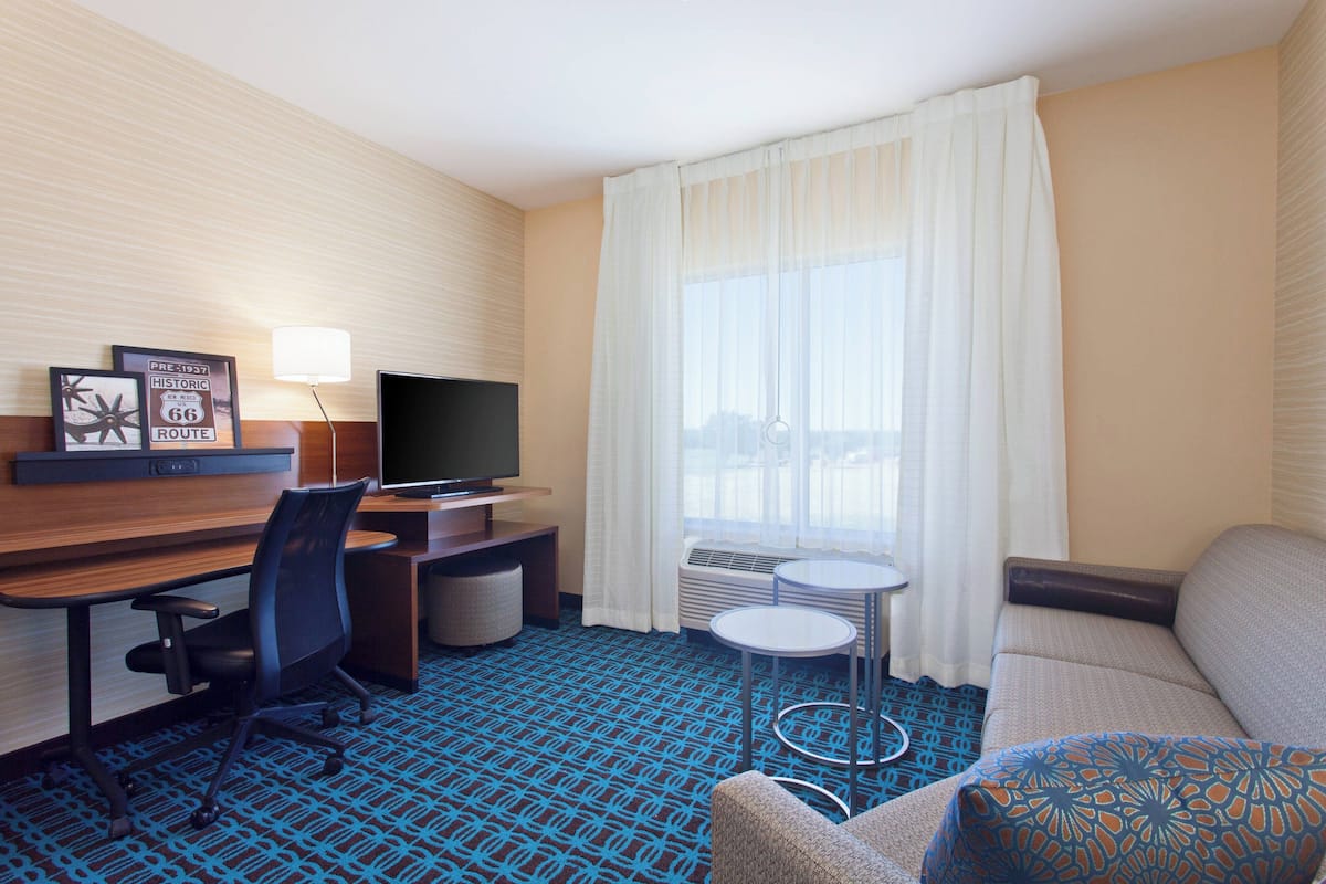 A hotel room with a desk, chair, television, sofa, and a view of the city through large windows.