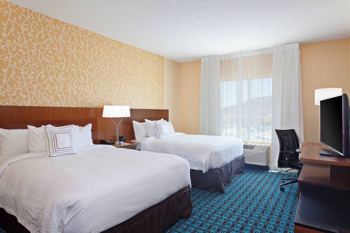 A hotel room with two beds, a desk with a television, and a large window with sheer curtains.