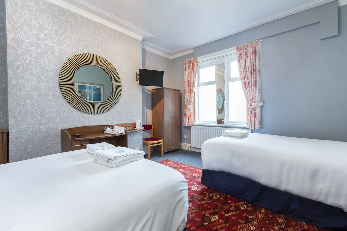 Quarto de hotel com duas camas, uma escrivaninha de madeira, um espelho, uma televisão e uma janela com cortinas.