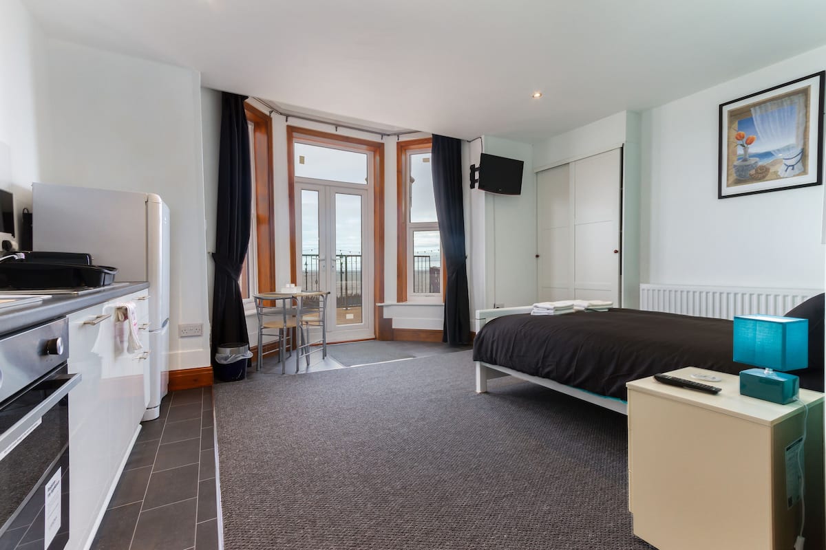 Quarto de hotel com uma cama, uma escrivaninha com um computador, uma televisão na parede e uma varanda com vista.