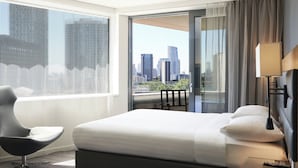 Ein Hotelzimmer mit einem großen Bett, einem modernen Sessel, einem Nachttisch und Blick auf eine Stadtlandschaft durch ein großes Fenster.