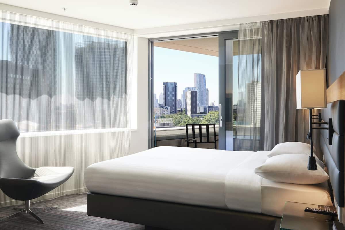 A hotel room with a large bed, a modern chair, a bedside table, and a view of a cityscape through a large window.