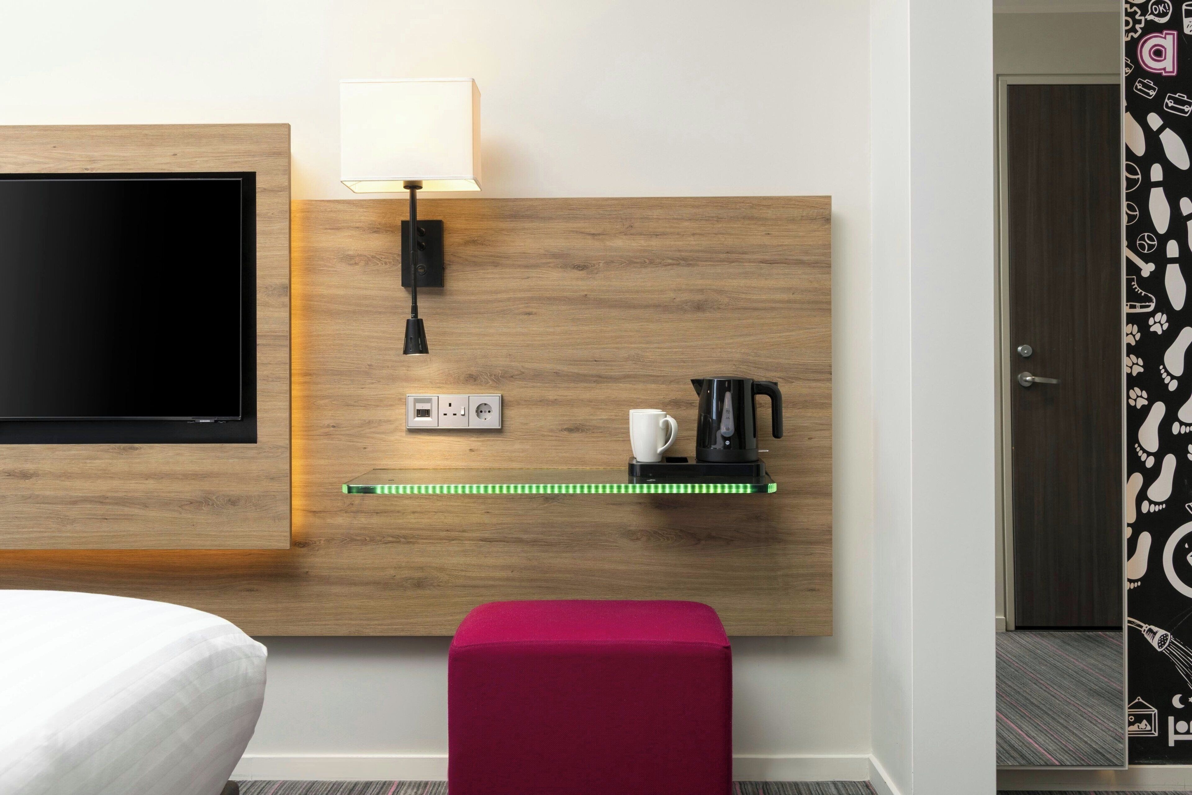 A hotel room with a flat-screen TV, a bedside lamp, a power outlet, a coffee maker, and a red ottoman.
