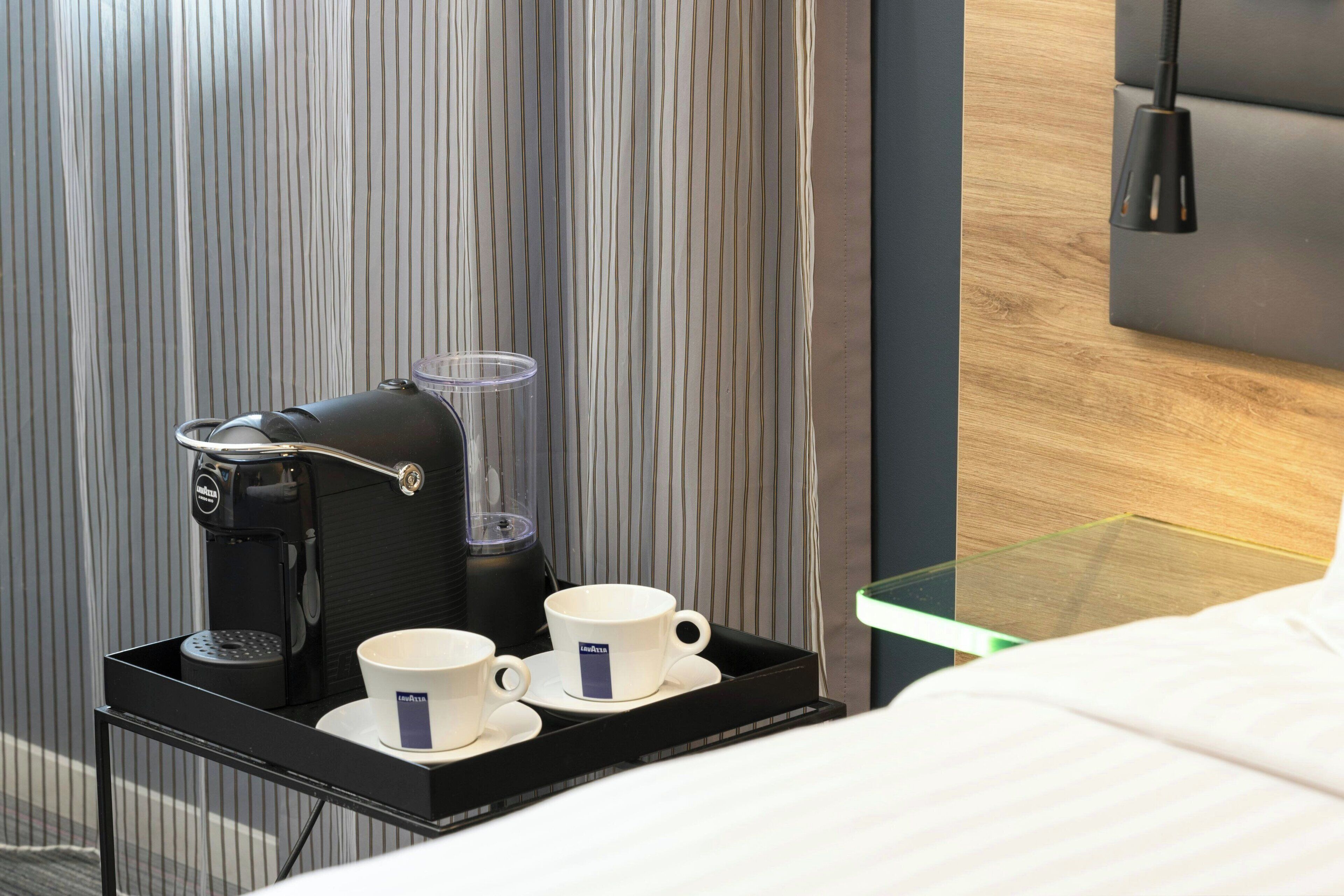 A hotel room with a bed, a bedside table with a coffee maker and cups, and a striped curtain.