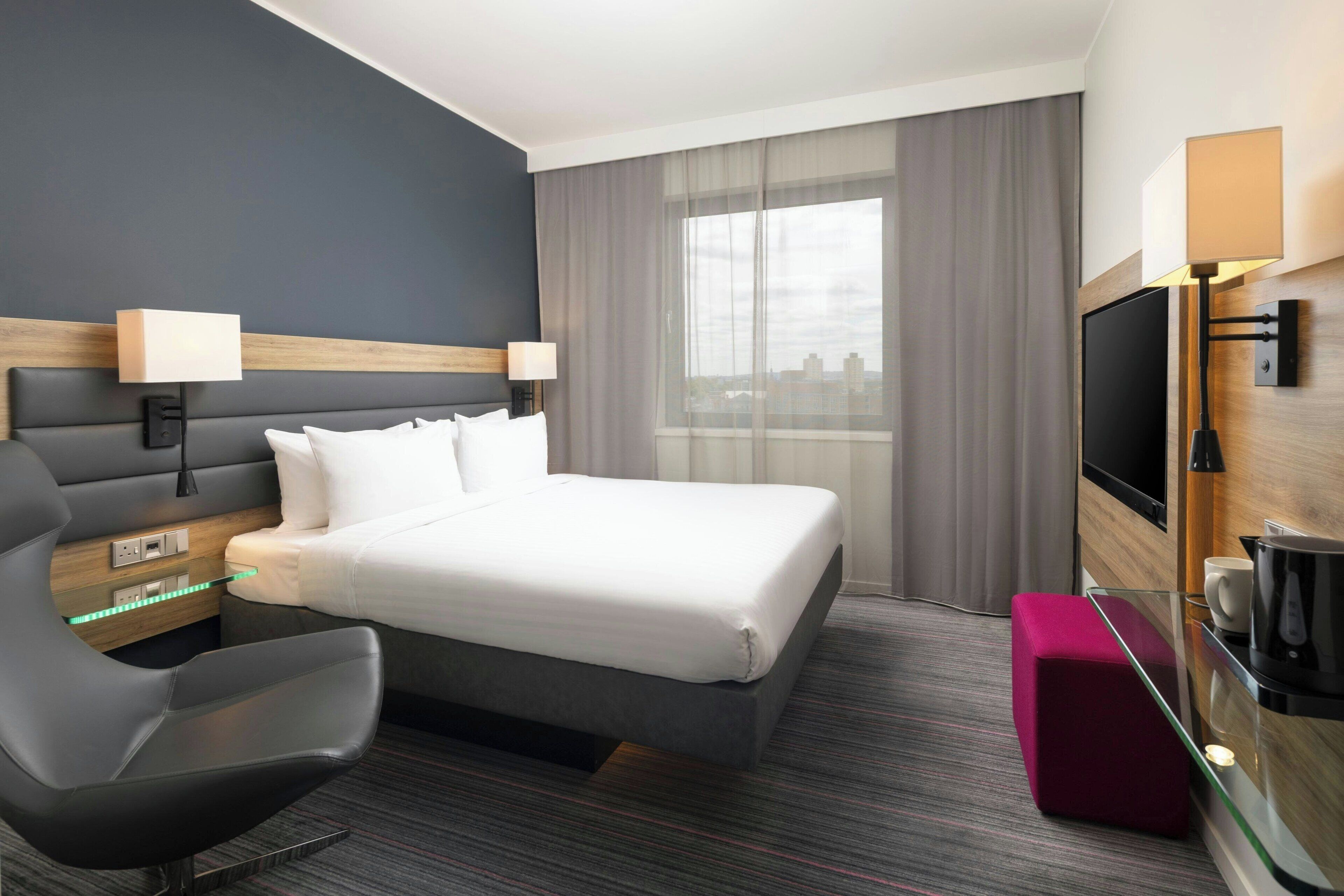 A modern hotel room with a large bed, a desk with a lamp, a TV, and a window with curtains.