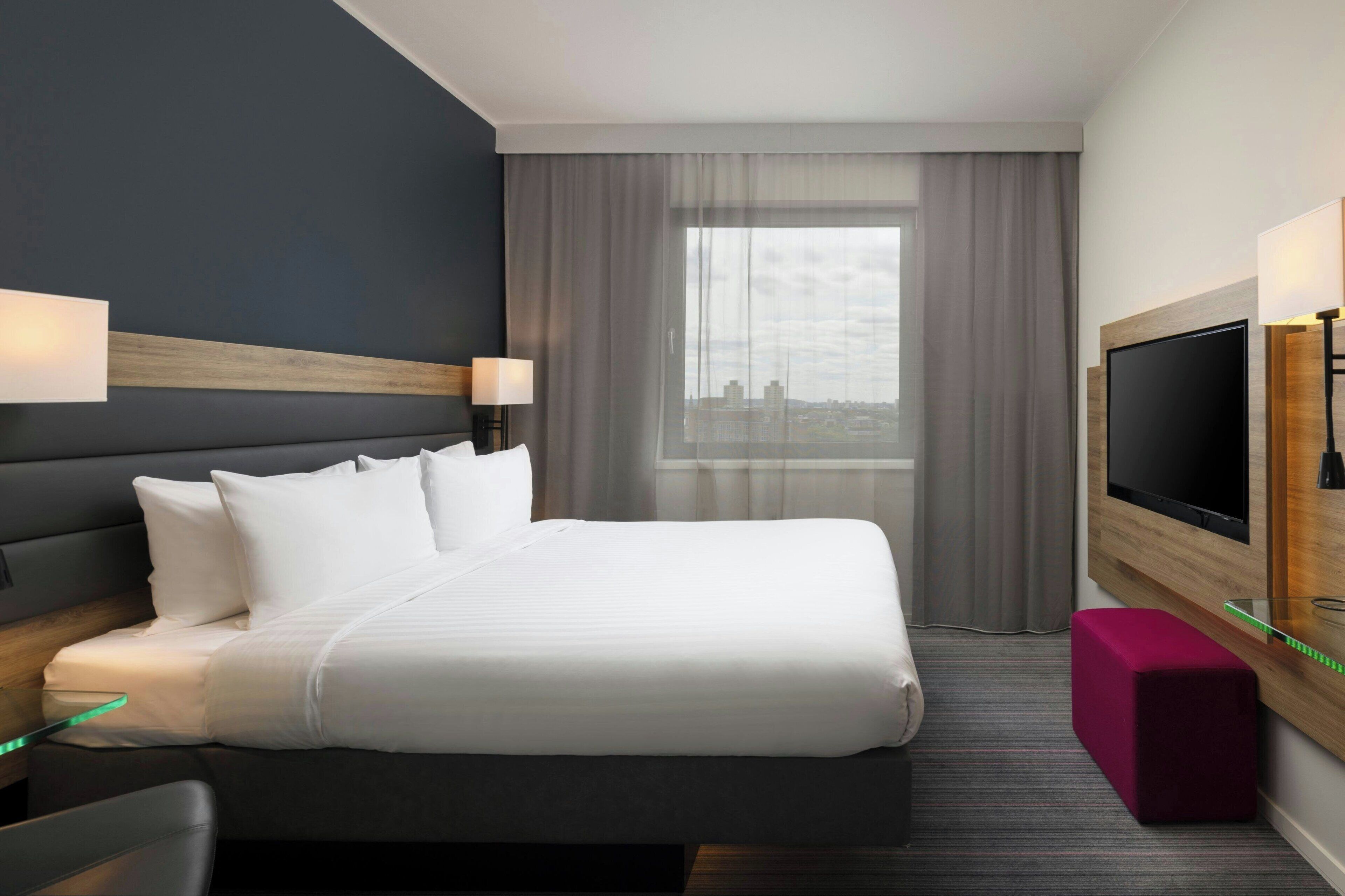 A modern hotel room with a large bed, a flat-screen TV, a desk with a chair, and a city view through the window.