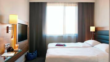 A hotel room with a bed, a desk with a lamp, a television, and a window with curtains.