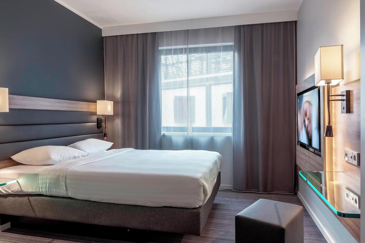 A modern hotel room with a large bed, a flat-screen TV mounted on the wall, and a window with curtains.
