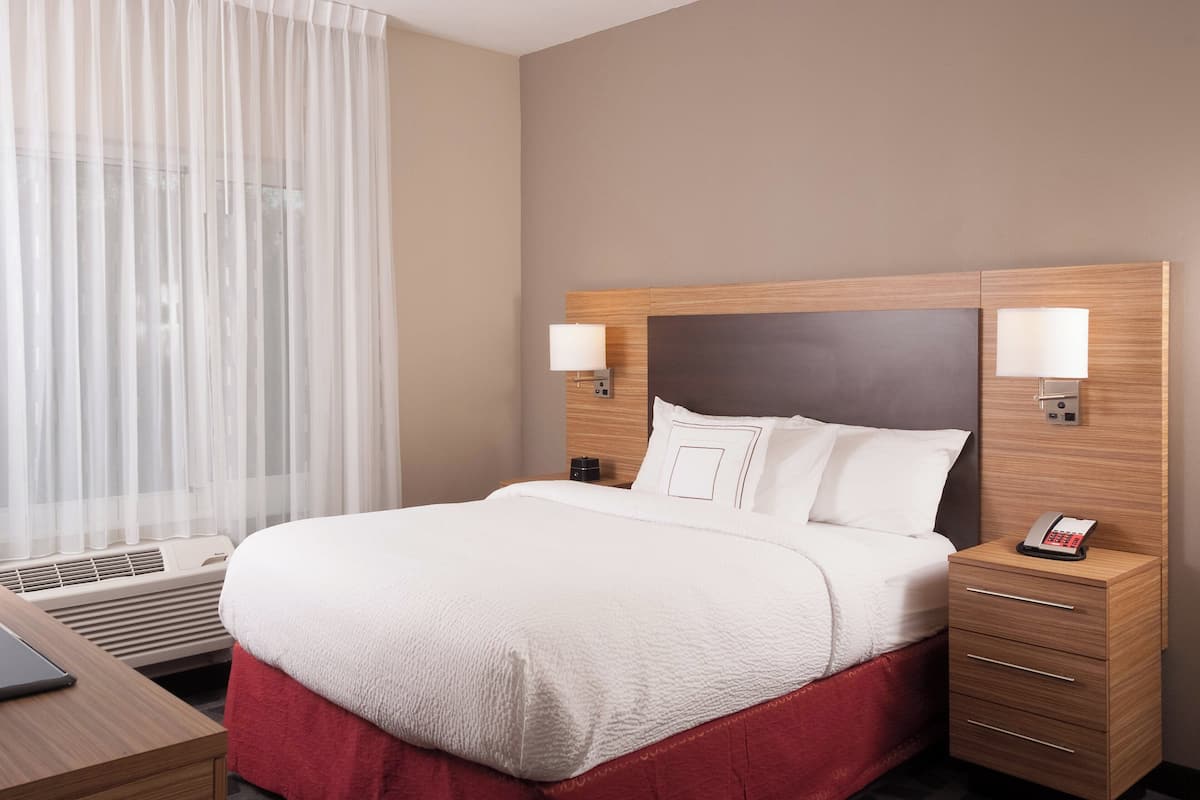 Une chambre d’hôtel avec un grand lit, une table de chevet avec une lampe, un téléphone et une fenêtre avec des rideaux légers.