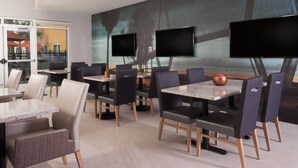 A modern dining area with multiple tables and chairs, large wall-mounted televisions, and a view of a bridge and palm trees.