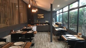 Uma área de refeições moderna com várias mesas e cadeiras, uma cozinha ao fundo e grandes janelas com vista para o verde.