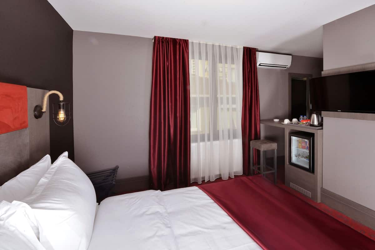 A hotel room with a bed, a desk with a TV, a minibar, and a window with curtains.