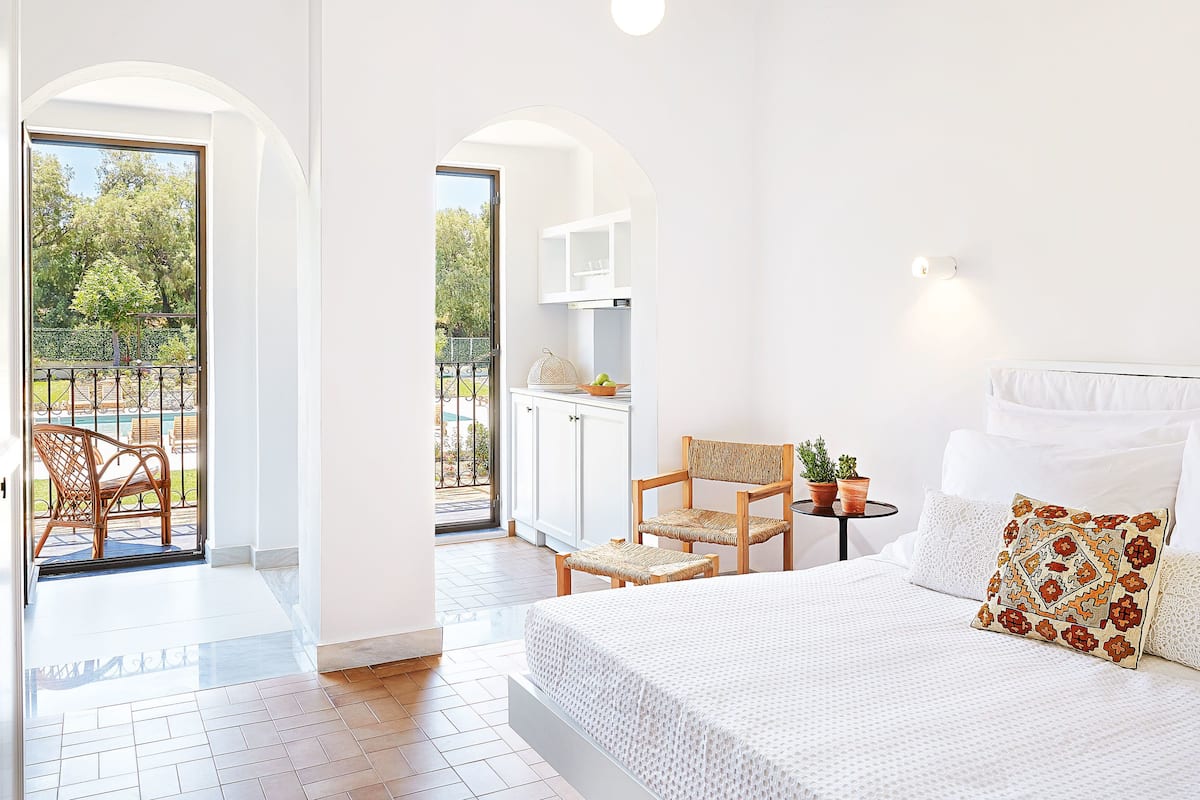 A spacious bedroom with a large bed, a dining area with chairs, and a view of a pool and garden.