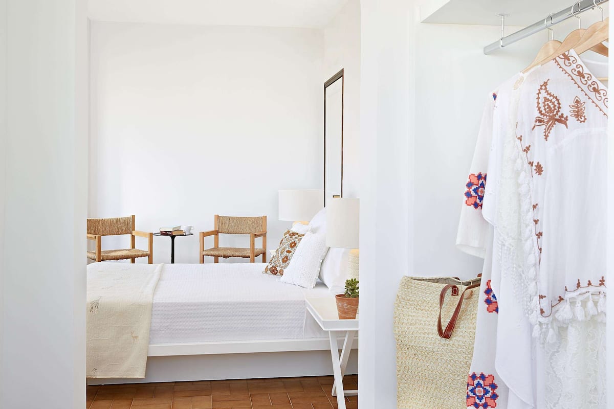 A bedroom with a bed, two chairs, a table, and a hanging garment rack.
