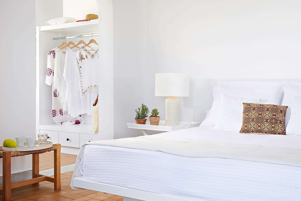 A bedroom with a bed, a bedside table with a lamp, a small table with a plant, and a wardrobe with clothes and a hat.