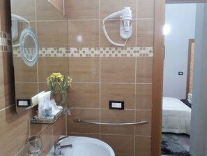 A bathroom with a white sink, a handheld showerhead, a glass shelf with toiletries, a mirror, and a bed in the adjacent room.