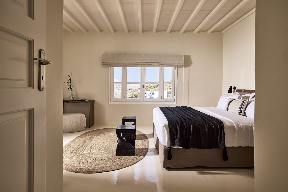 A bedroom with a bed, bedside table, lamp, and a view of the outdoors through a window.