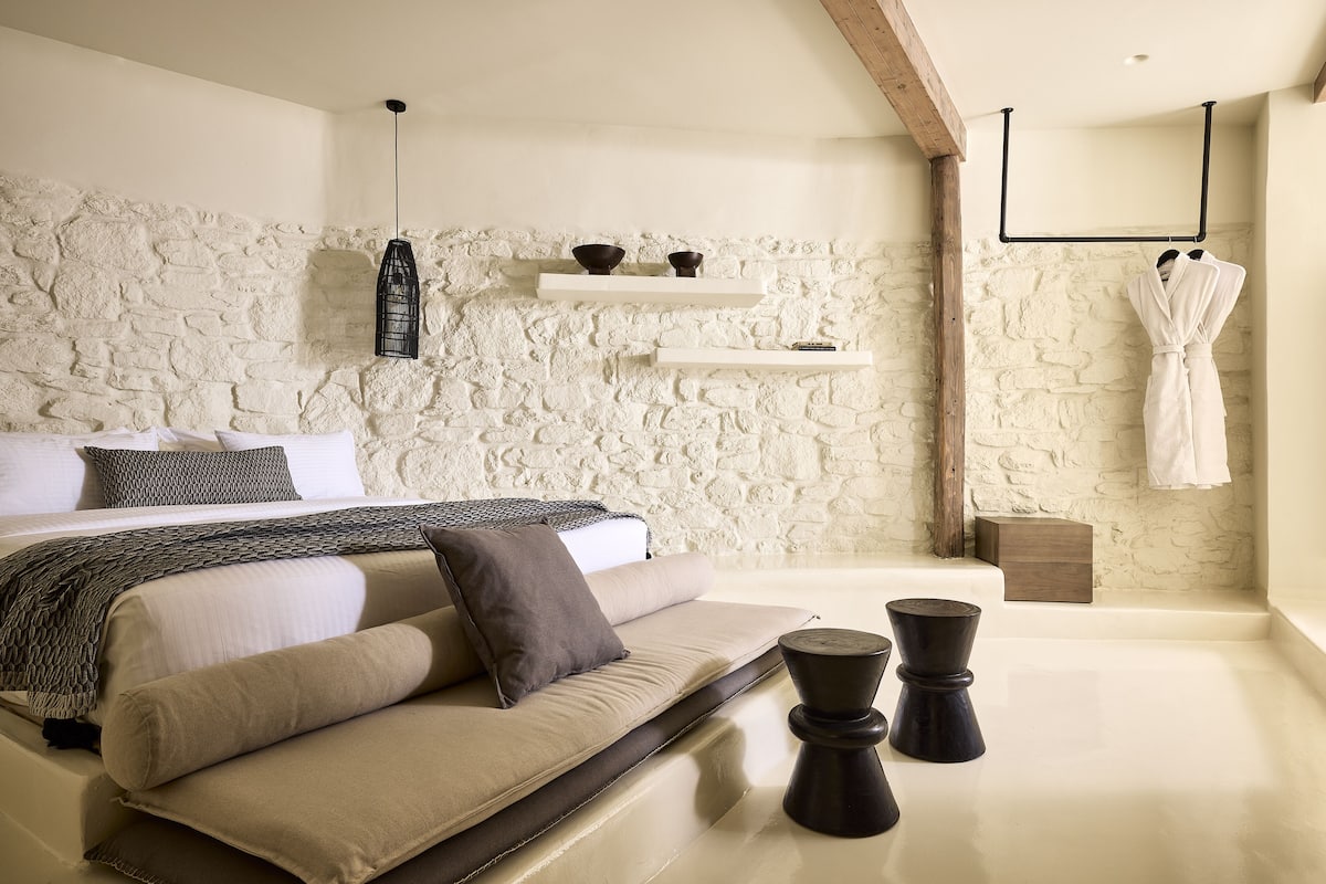 A modern bedroom with a stone wall, a bed with beige bedding, a hanging robe, and a small wooden nightstand.
