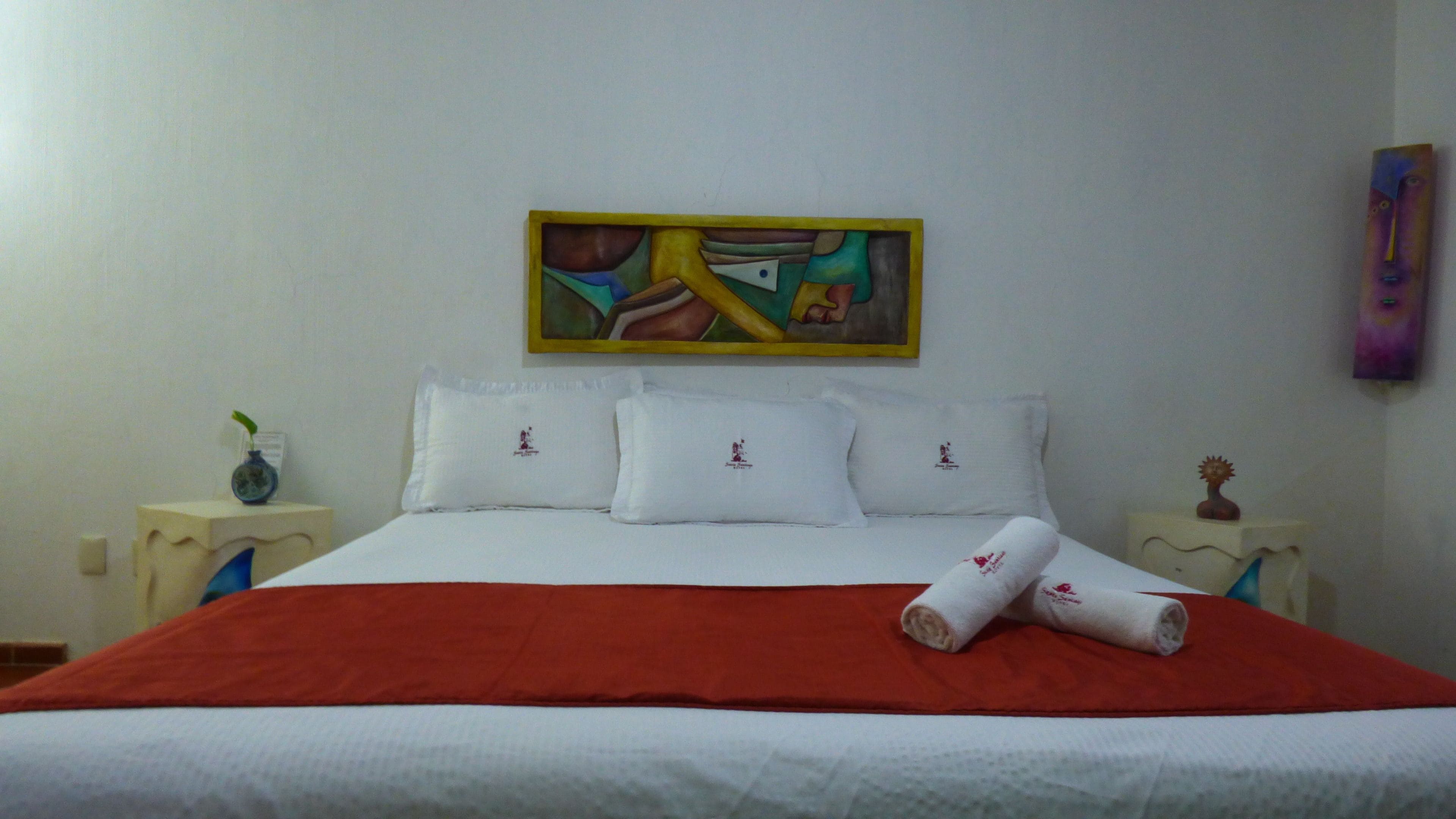 A hotel room with a bed, white pillows, a red blanket, a painting on the wall, and a small vase on the nightstand.