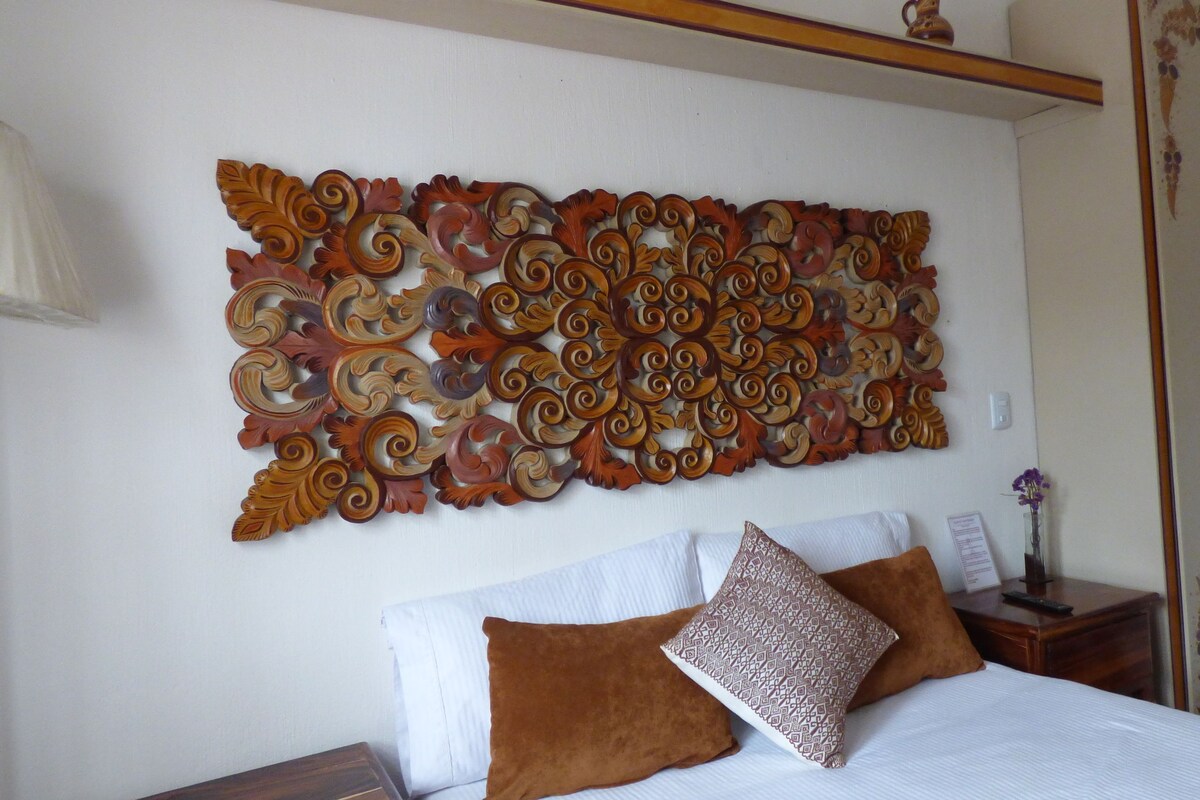 Une chambre à coucher avec un grand panneau mural décoratif, un lit avec une literie blanche et deux oreillers marron, une table de chevet avec un vase et une télécommande, et une tête de lit en bois.