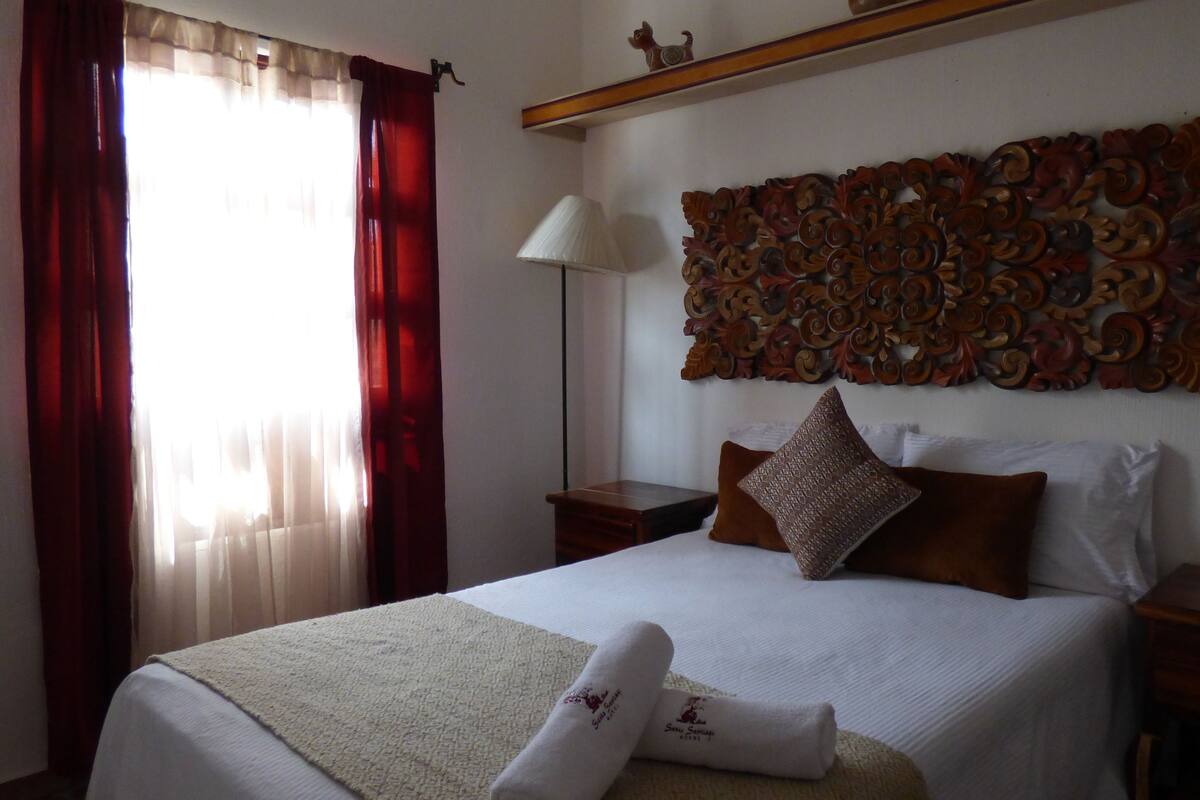 A hotel room with a large bed, a decorative wall panel, a nightstand, a lamp, and a window with curtains.