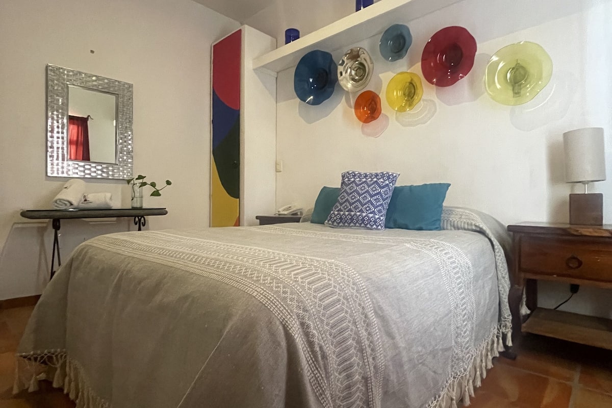 A bedroom with a bed, a bedside table, a mirror, and a wall with colorful glass plates.