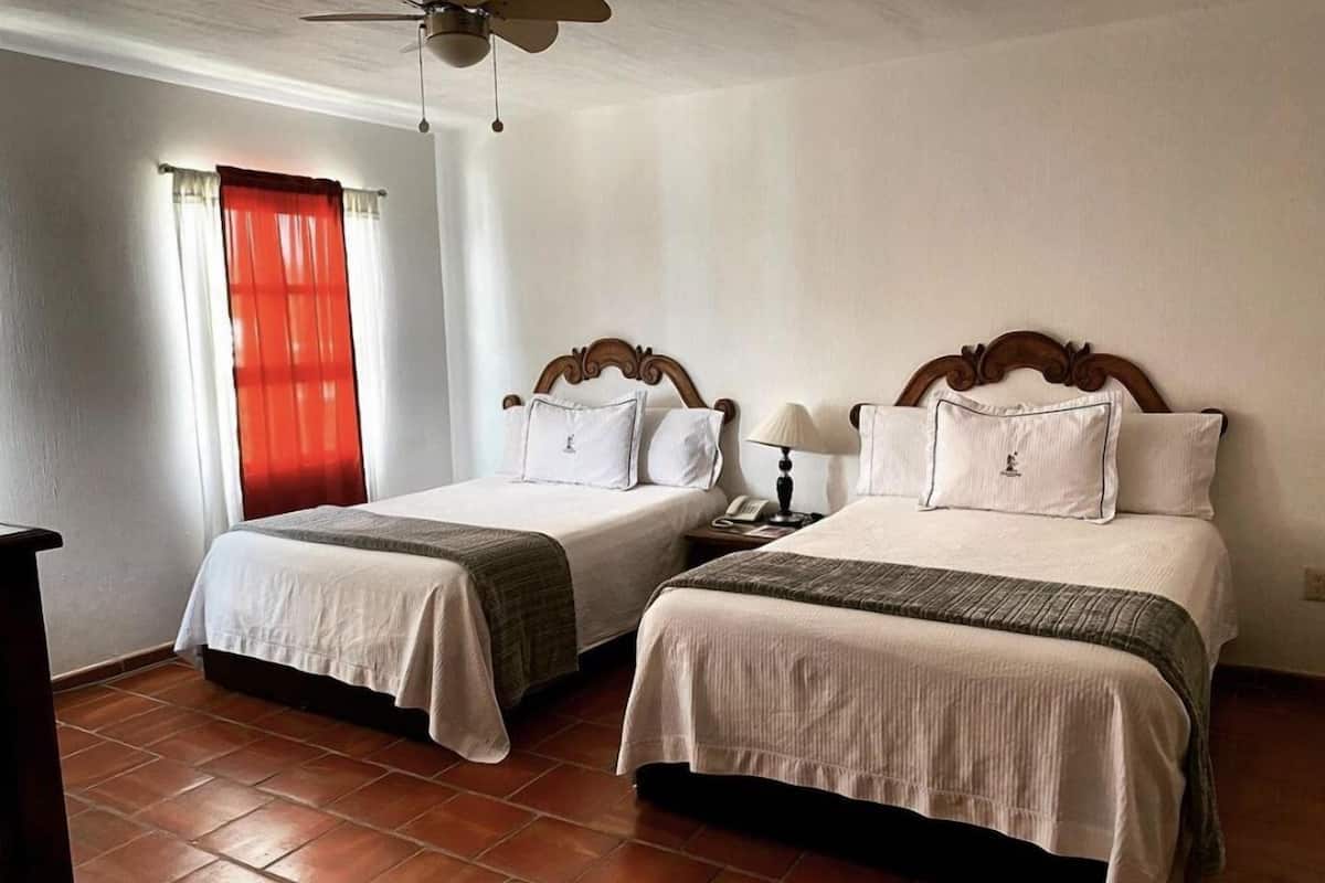 A hotel room with two beds, a ceiling fan, a window with red curtains, and a tiled floor.
