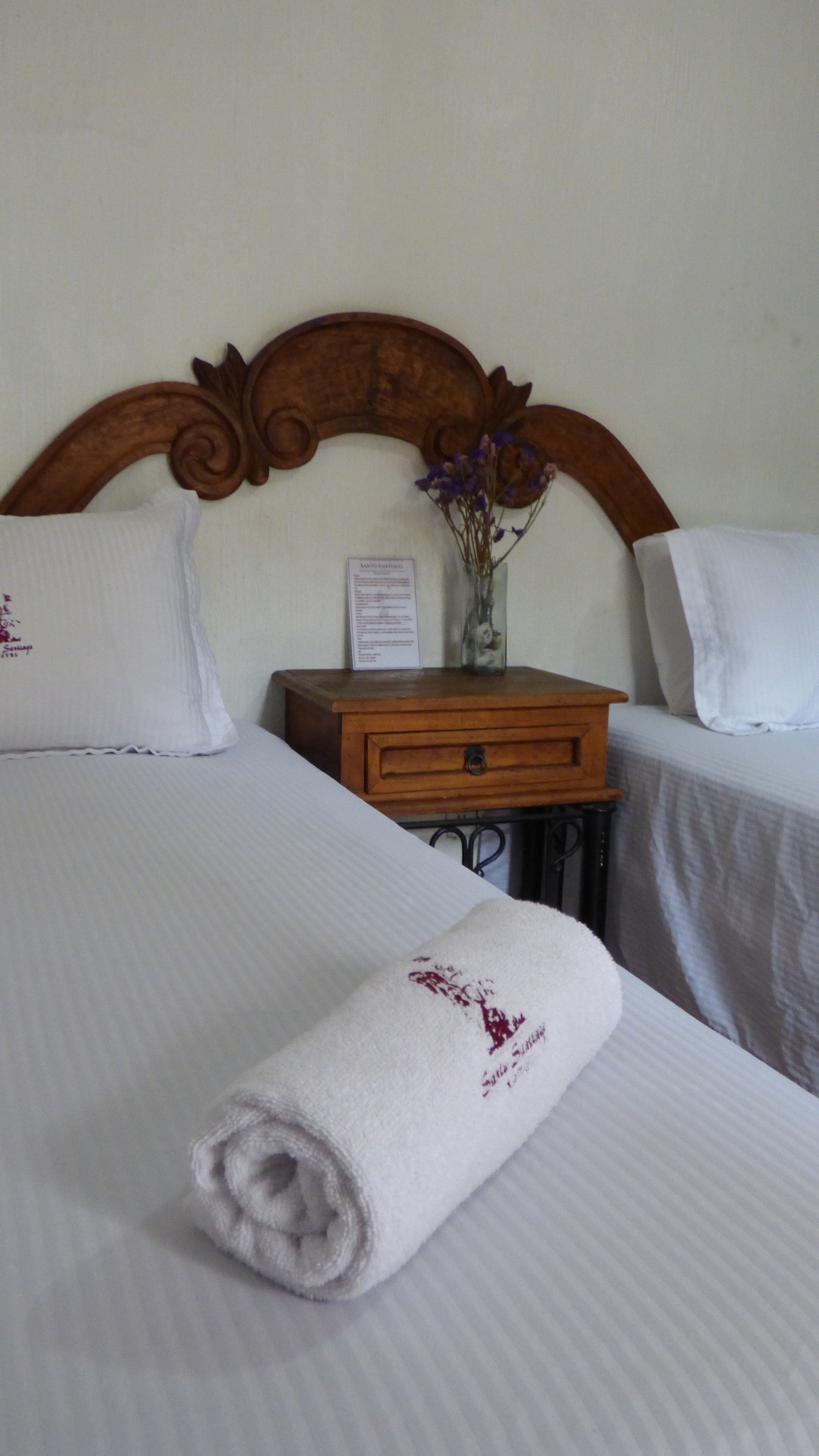 A neatly made bed with a rolled towel, a wooden nightstand with a vase of flowers, and two pillows.