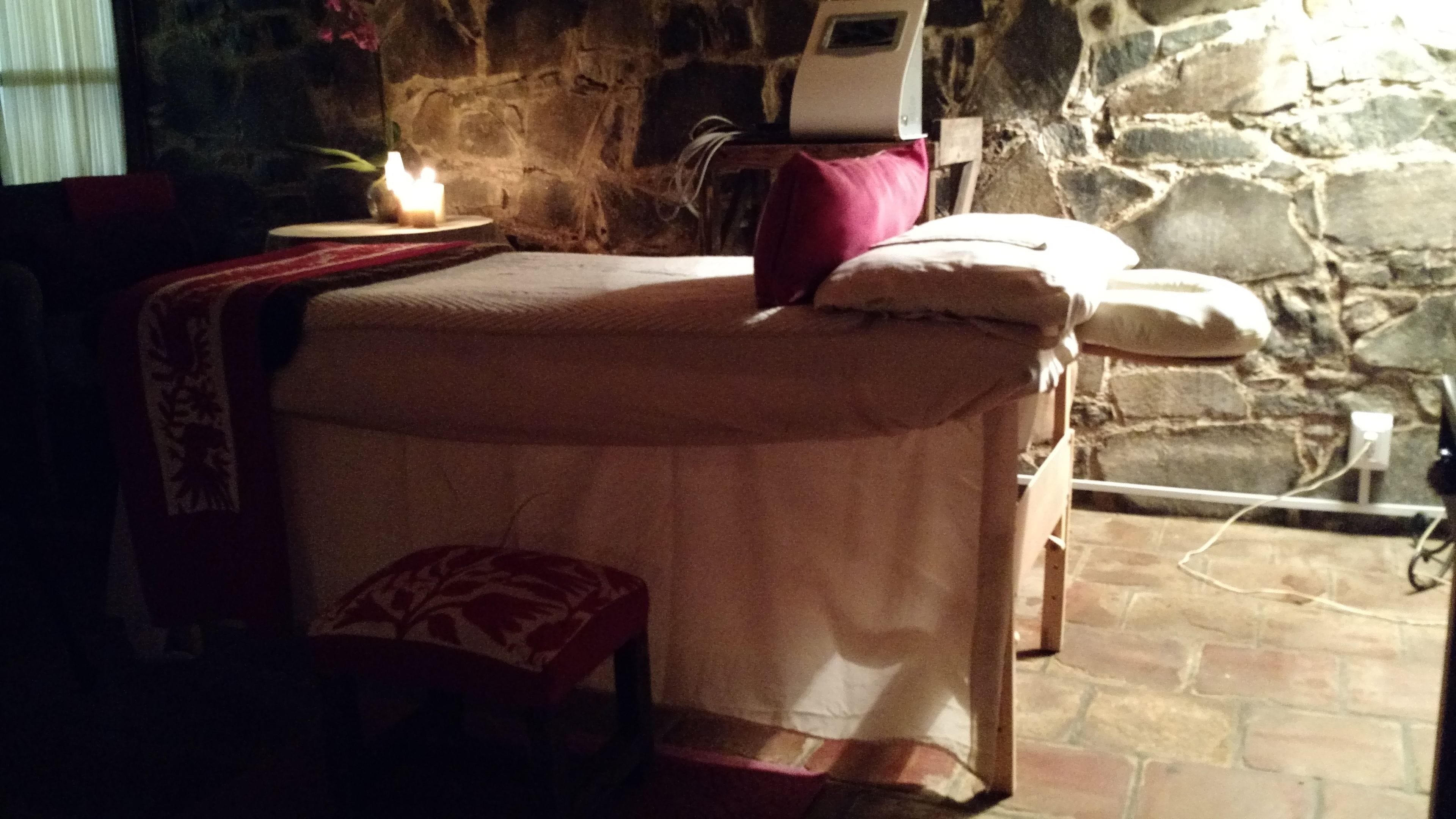 Una habitación de paredes de piedra con una cama, una mesita con una vela y suelo de piedra.