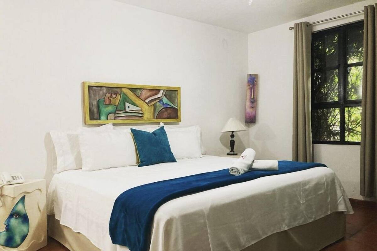 A hotel room with a bed, a painting on the wall, a lamp, and a window with curtains.