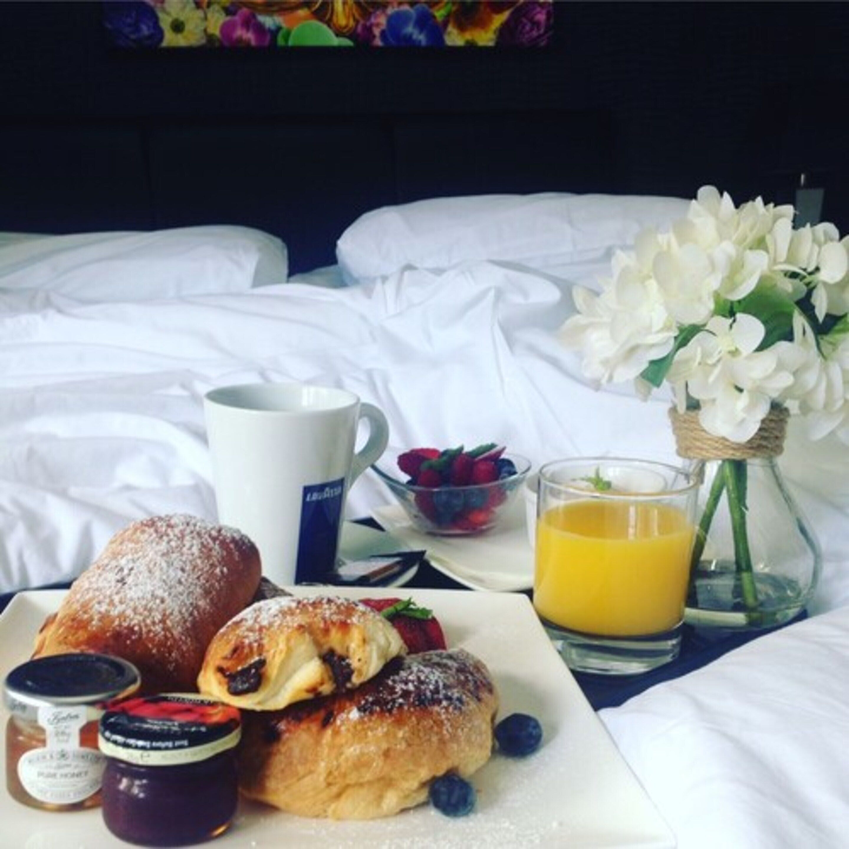 A neatly made bed with white linens, a plate of pastries, a cup of coffee, a glass of orange juice, and a vase with white flowers.