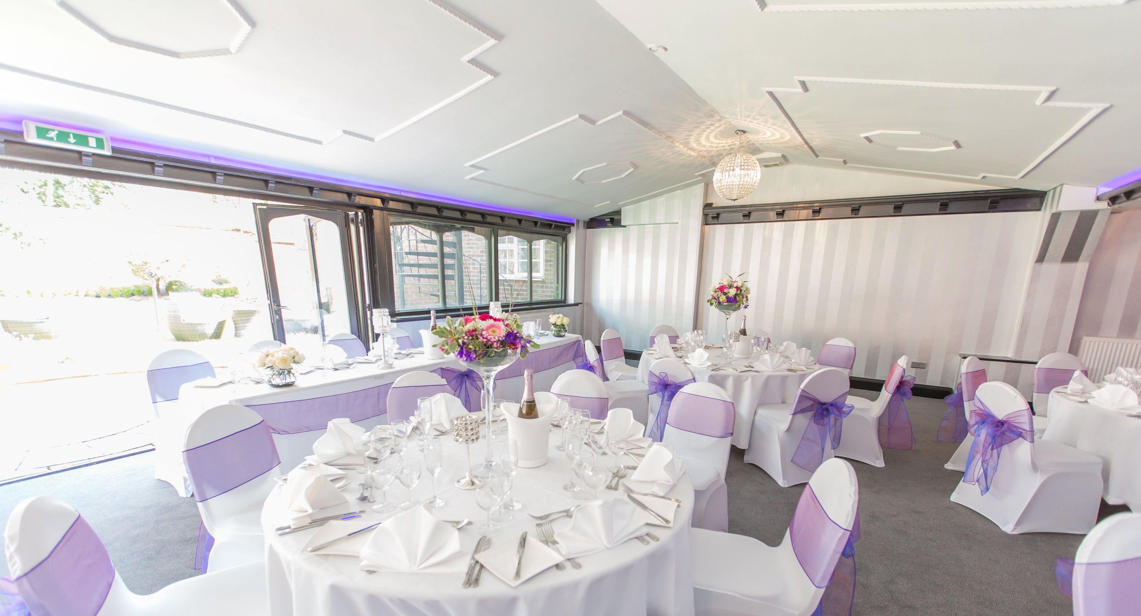 Un salón de banquetes con mesas redondas dispuestas para un evento formal, con manteles blancos, lazos morados en las sillas y centros de mesa florales.
