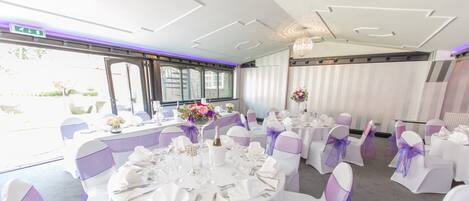 Un salón de banquetes con mesas redondas dispuestas para un evento formal, con manteles blancos, lazos morados en las sillas y centros de mesa florales.
