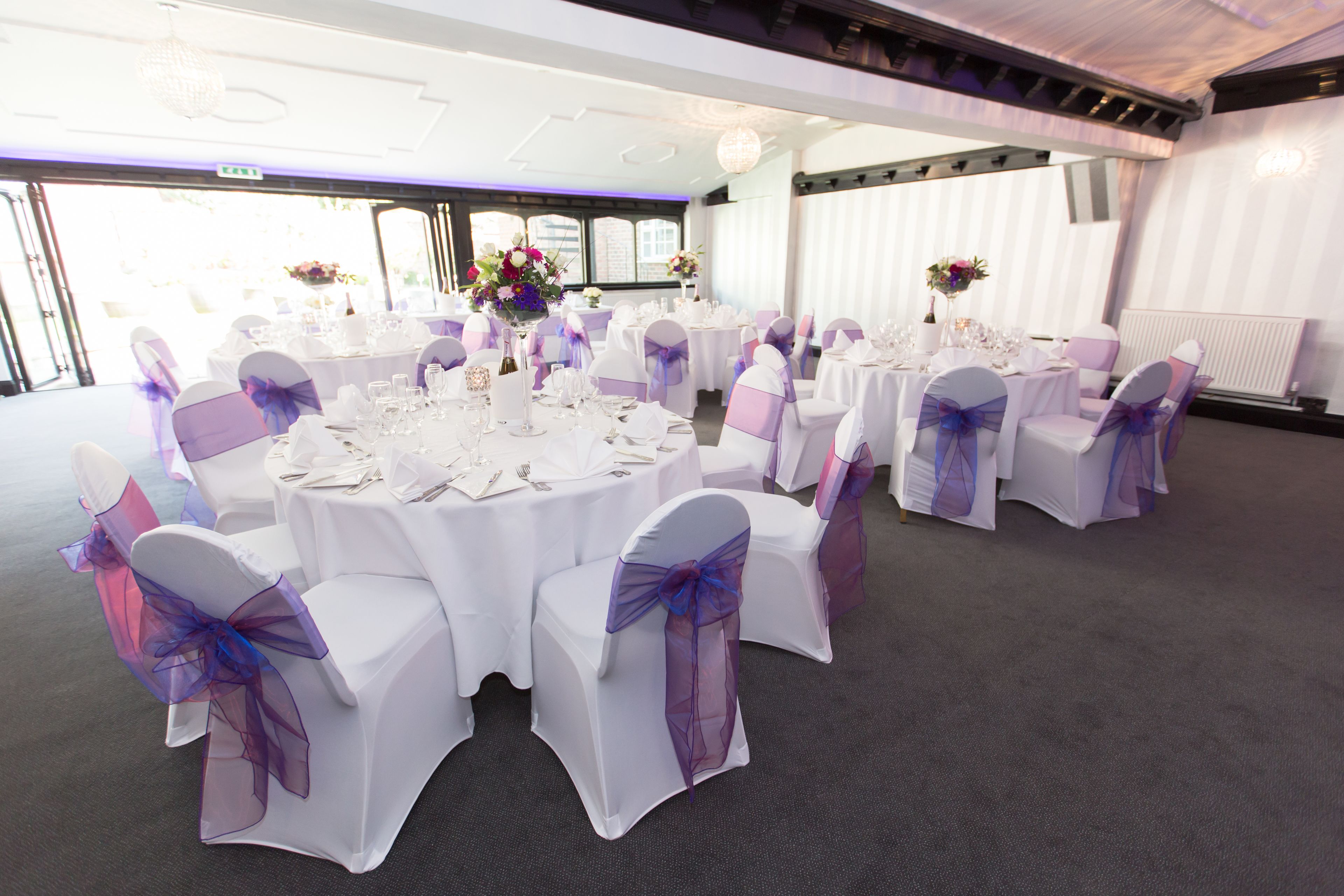 A banquet hall with round tables set for dining, white chairs with purple sashes, and floral centerpieces.