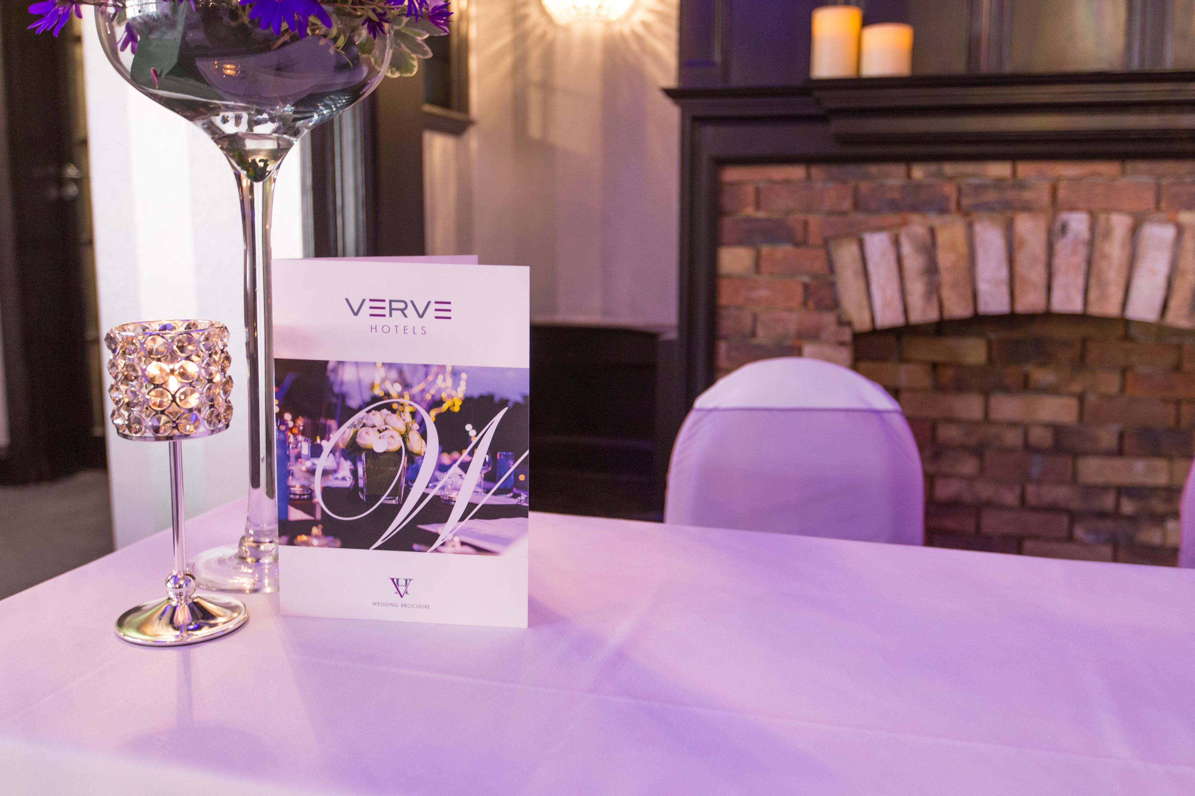 Una mesa con un folleto de Verve Hotels, un florero de cristal decorativo y un candelabro.