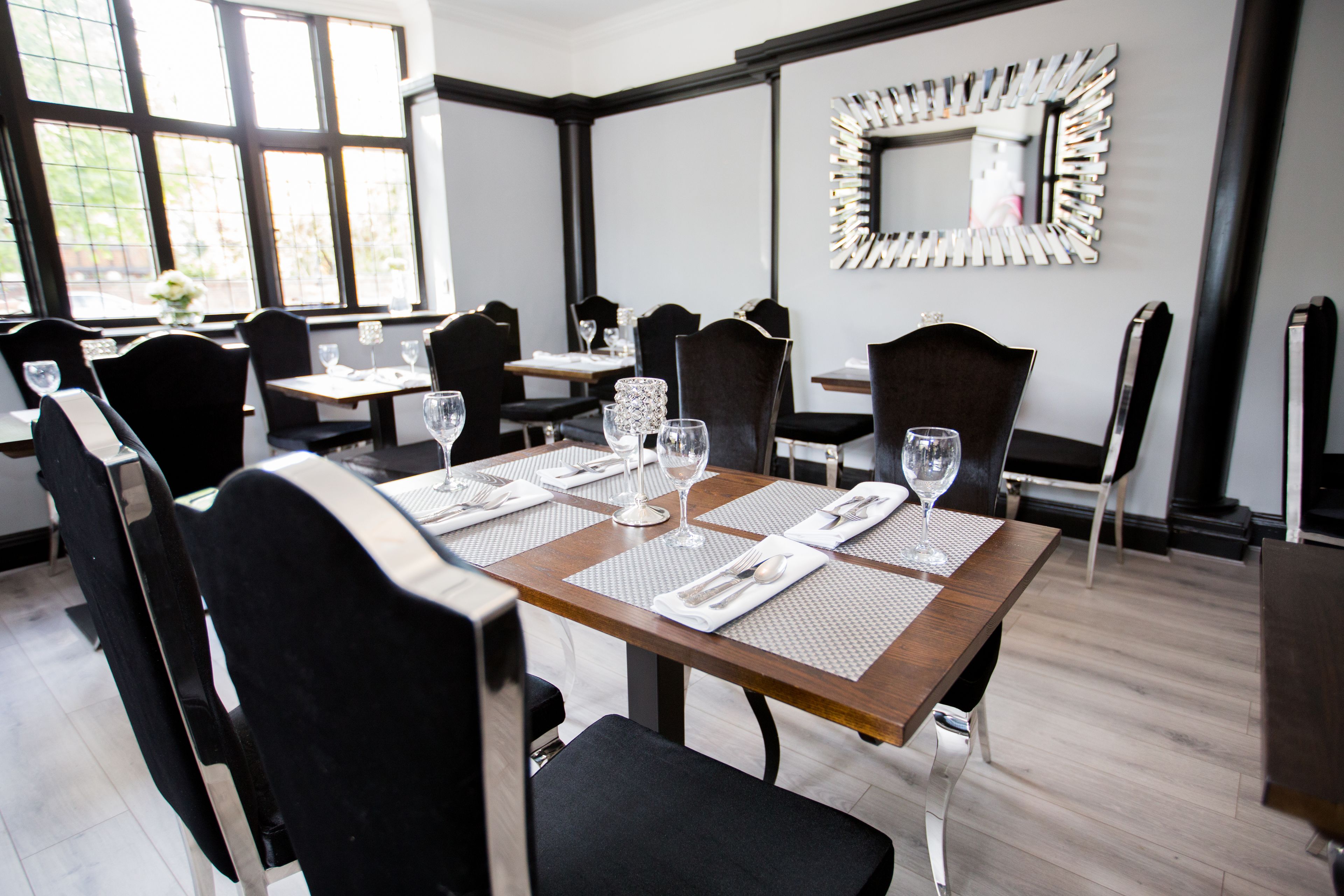Un coin repas avec des tables dressées pour quatre personnes, chacune agrémentée d’un set de table, de couverts et de verres à vin. La pièce présente un design moderne, avec des chaises noires et un grand miroir au mur.