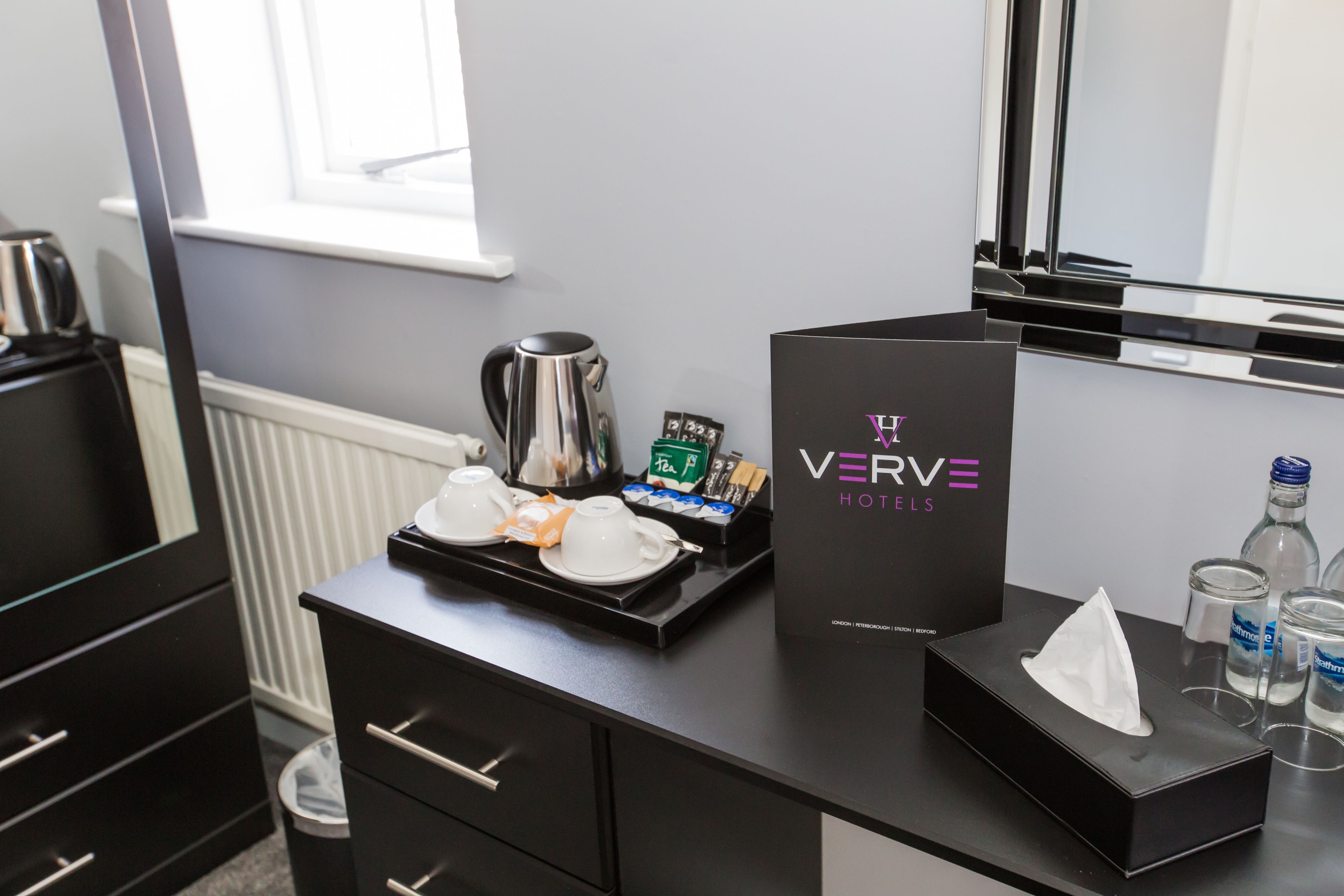 Quarto de hotel com minibar preto, bandeja com chá e biscoitos, caixa de lenços e uma placa VERVE Hotels.