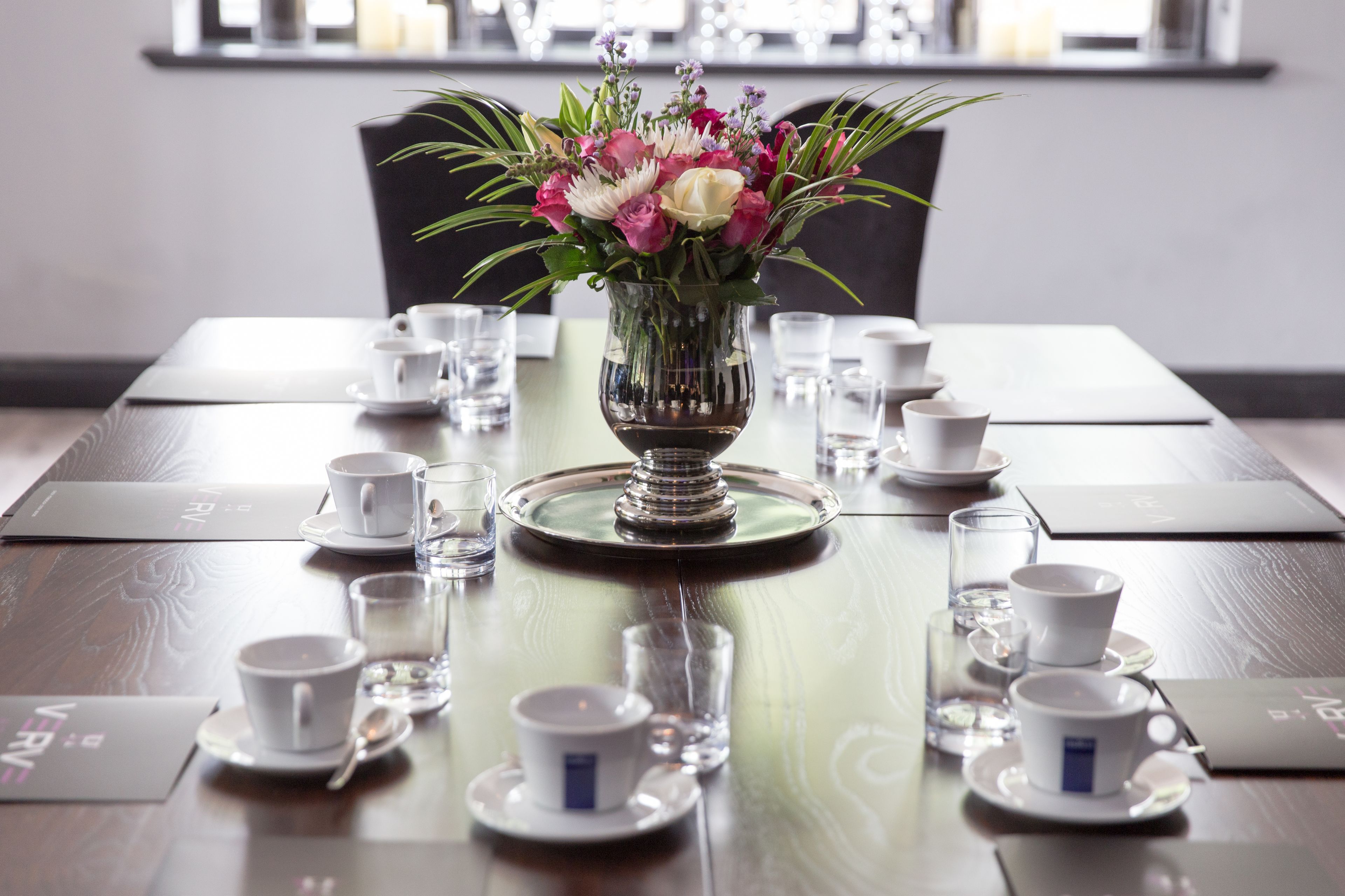 Una mesa de comedor dispuesta con tazas y platillos blancos, una copa de cristal transparente y un florero con flores.