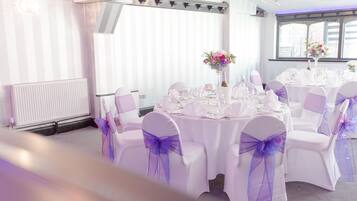 Un salón de banquetes con mesas redondas dispuestas para un evento formal, sillas blancas con lazos morados y centros de mesa florales.