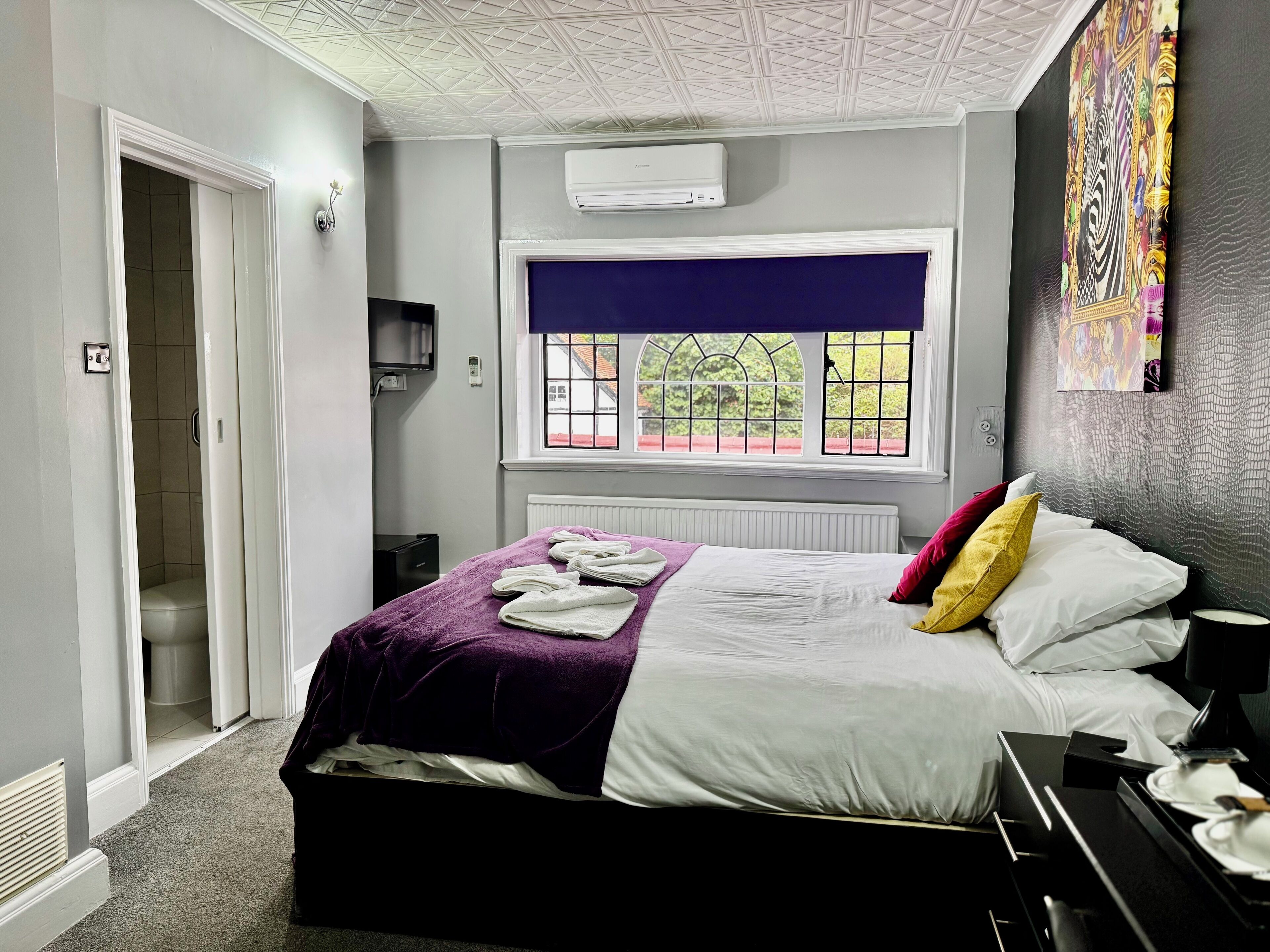 Habitación de hotel ordenada con cama, ventana con vistas, radiador y aire acondicionado instalado en la pared.