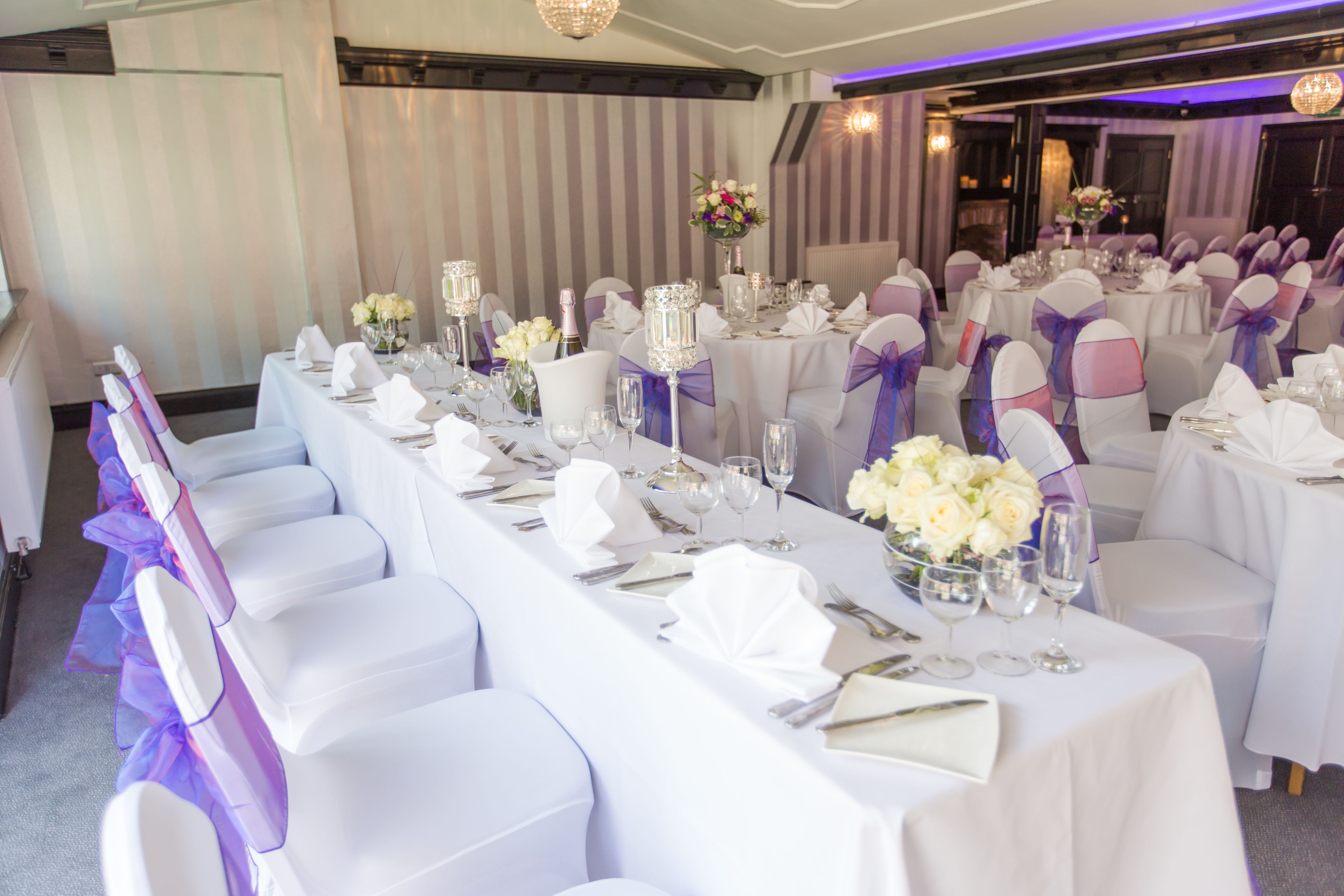 A banquet hall with long tables set for a formal event, featuring white tablecloths, neatly arranged silverware, and floral centerpieces.
