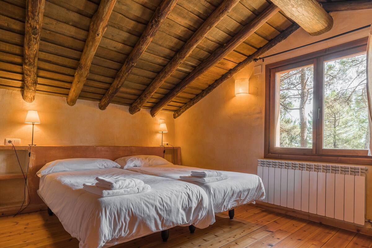 A cozy bedroom with a wooden ceiling, a large window with a view of trees, and two bedside lamps.