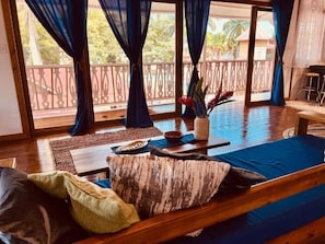 A room with a wooden floor, a sofa with cushions, a small table with a bowl and a vase, and a view of a balcony with a railing and trees. - Casa Le Parc (Bocas del Toro)