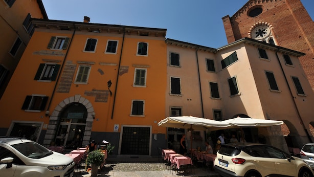 A building with a clock tower, outdoor seating area, and parked cars. - Casa Sottoriva (Verona)