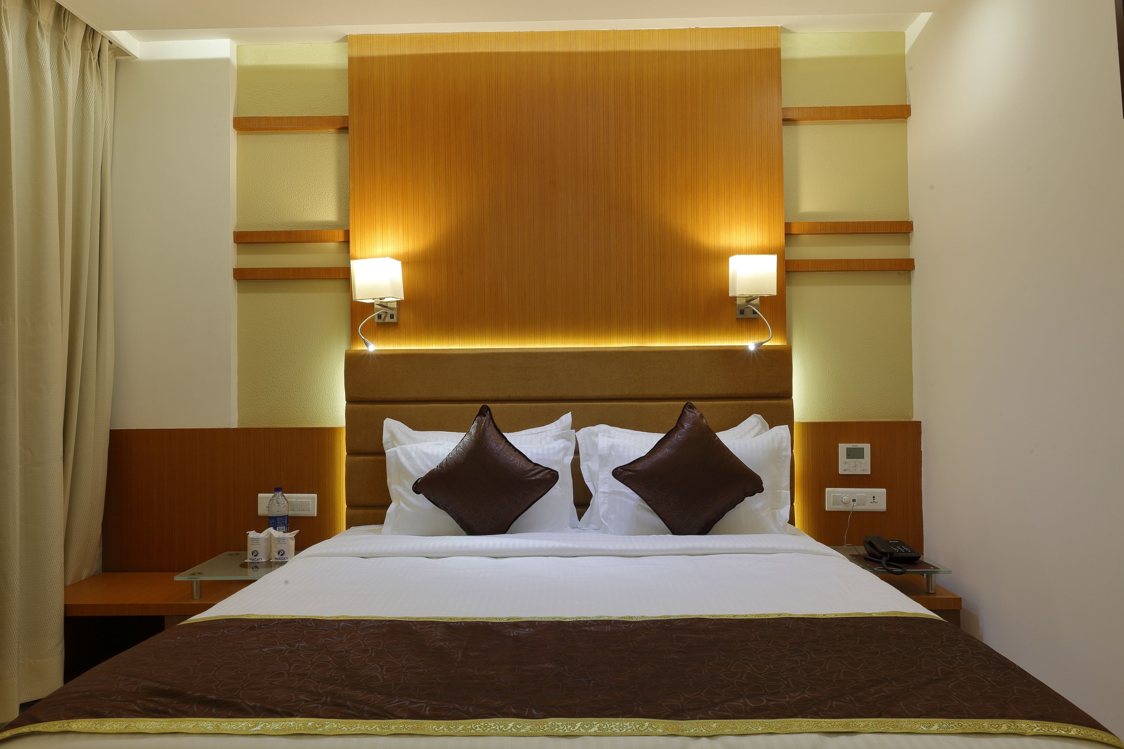 A hotel room with a large bed, two bedside lamps, a nightstand with a phone, and a wall-mounted control panel.