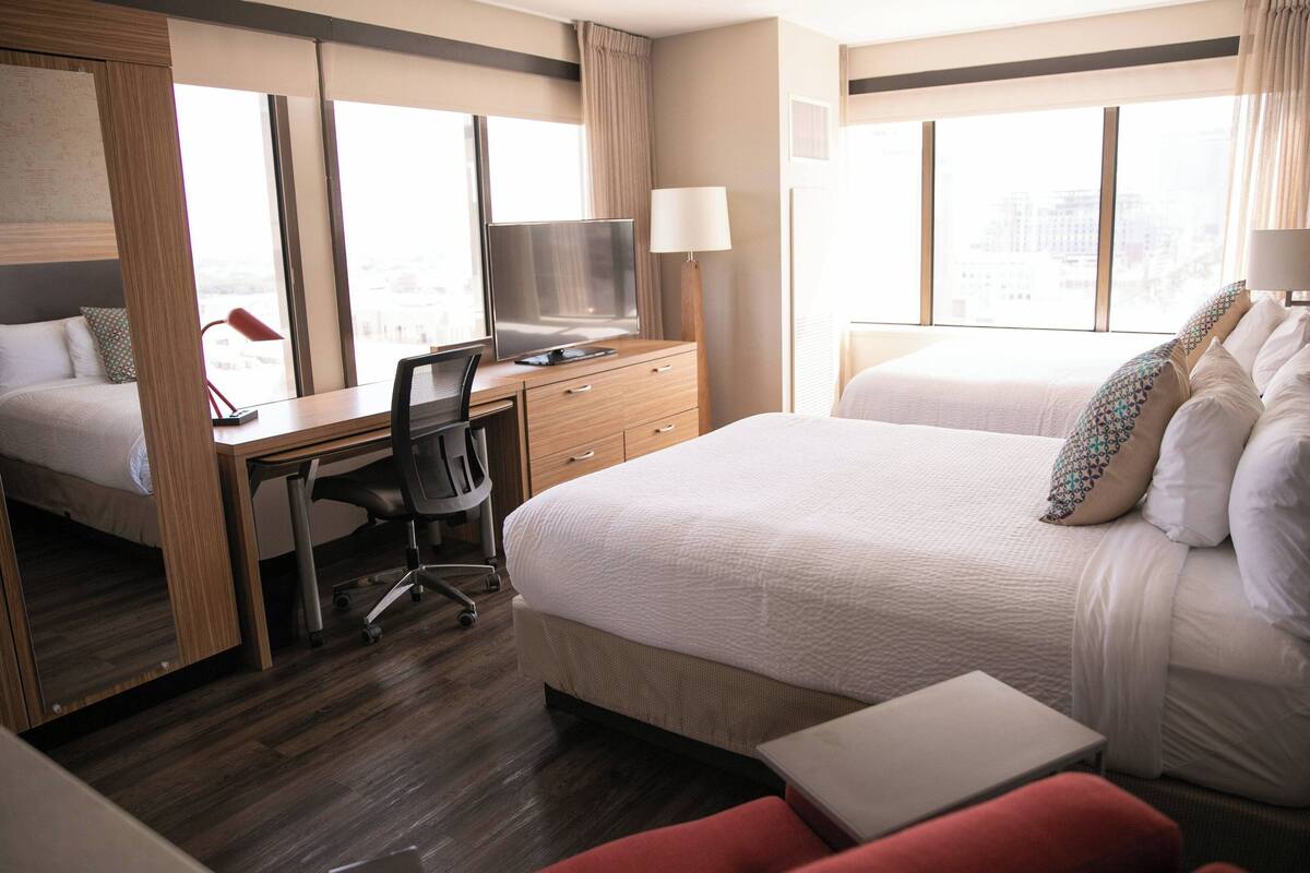 A hotel room with a bed, desk, chair, TV, and windows.