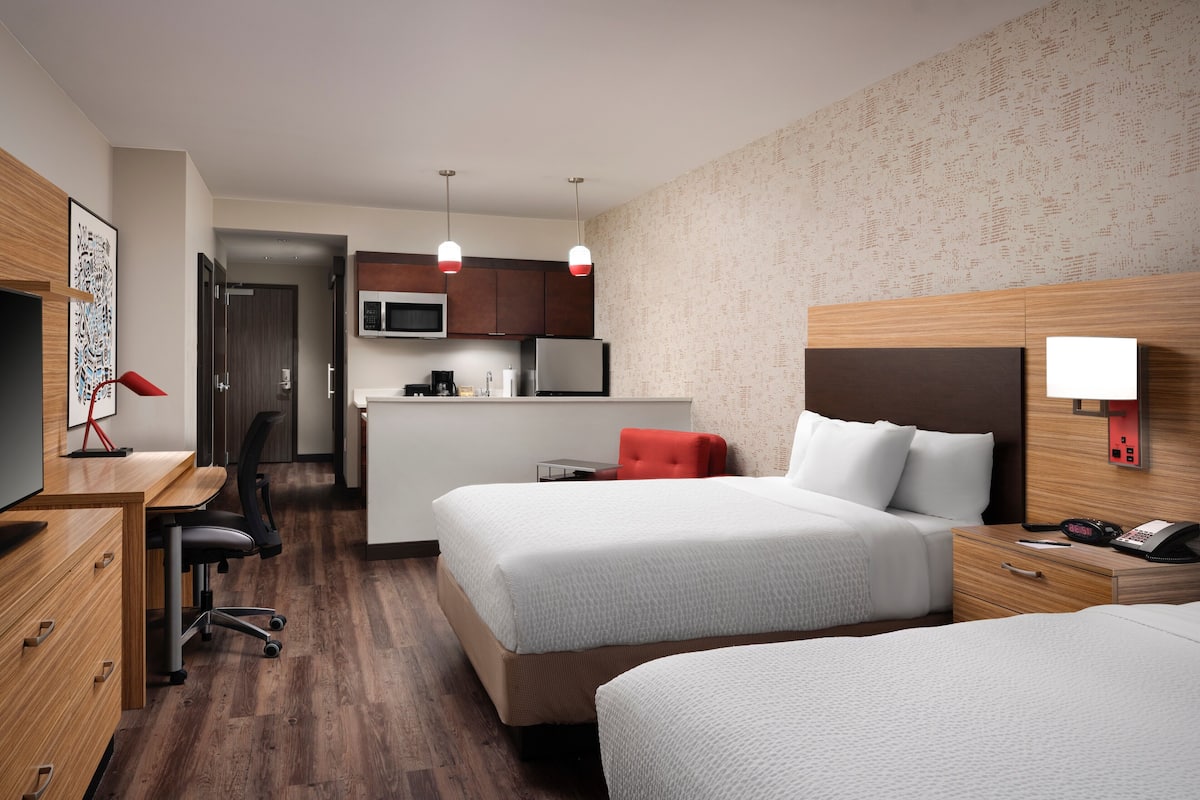 A hotel room with two beds, a desk, a chair, a microwave, a coffee maker, and a small kitchenette.