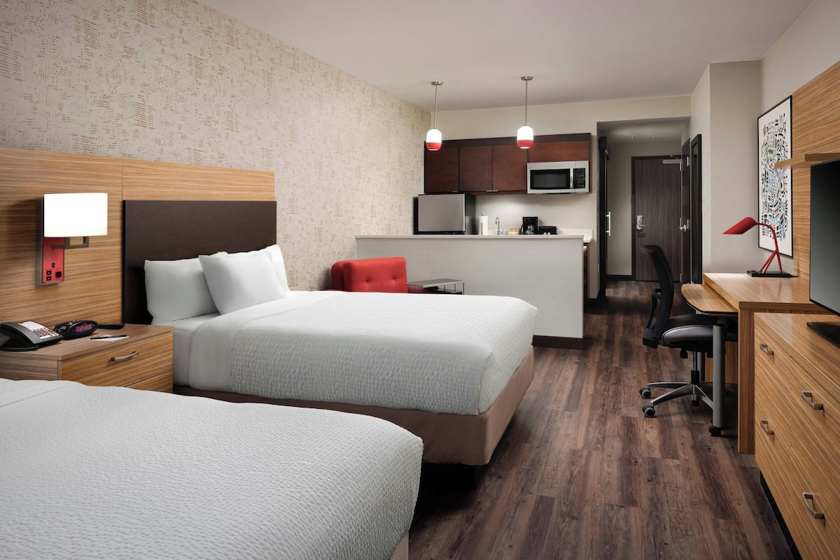 A hotel room with two beds, a desk, a chair, a microwave, a small kitchenette, and a door leading to another room.