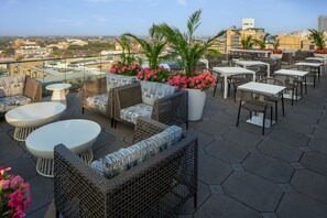 Eine Dachterrasse mit Korbmöbeln, weißen Tischen und Topfpflanzen.